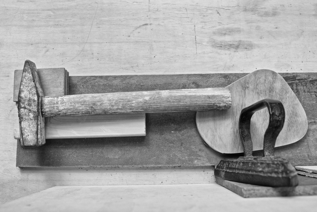 Black and white photograph of a hammer with weathered metal head and wooden handle resting on a steel workbench with iron tools