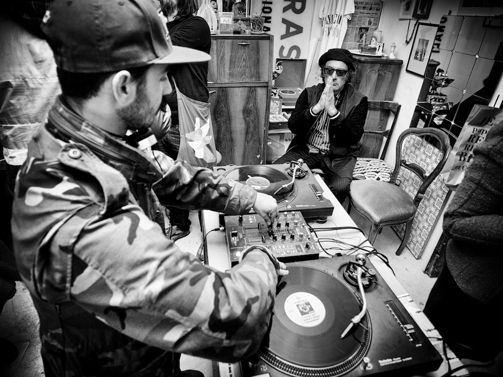 Black and white photograph of DJs working with turntables and mixing equipment in an intimate studio setting.