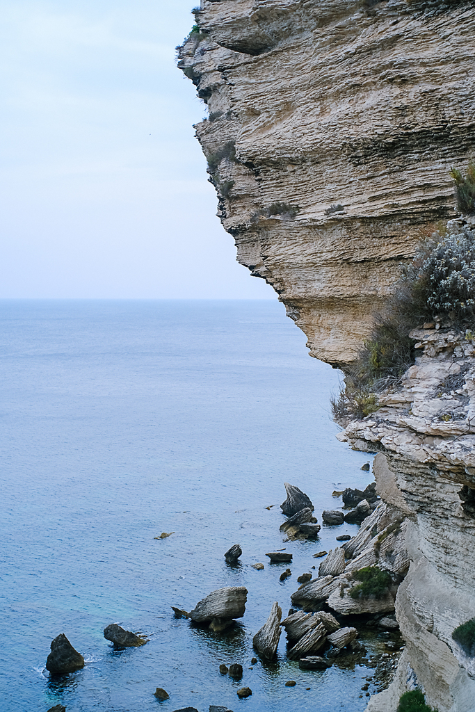 Dramatic rocky coastal cliff face overlooking calm blue Mediterranean sea with scattered rocks below, part of Wonderland series.