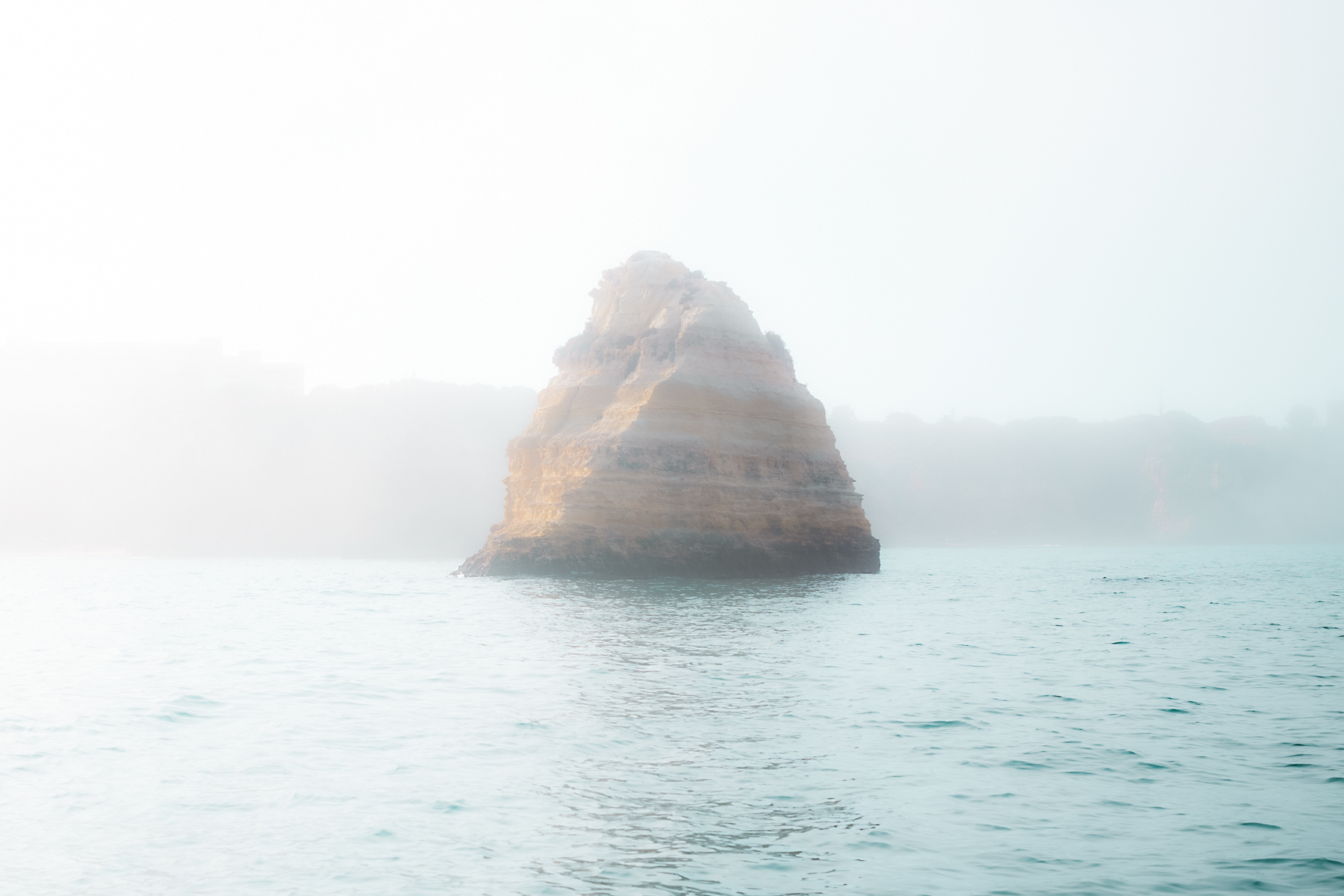 A solitary rocky island emerges from misty Atlantic waters, shrouded in fog under pale morning light.