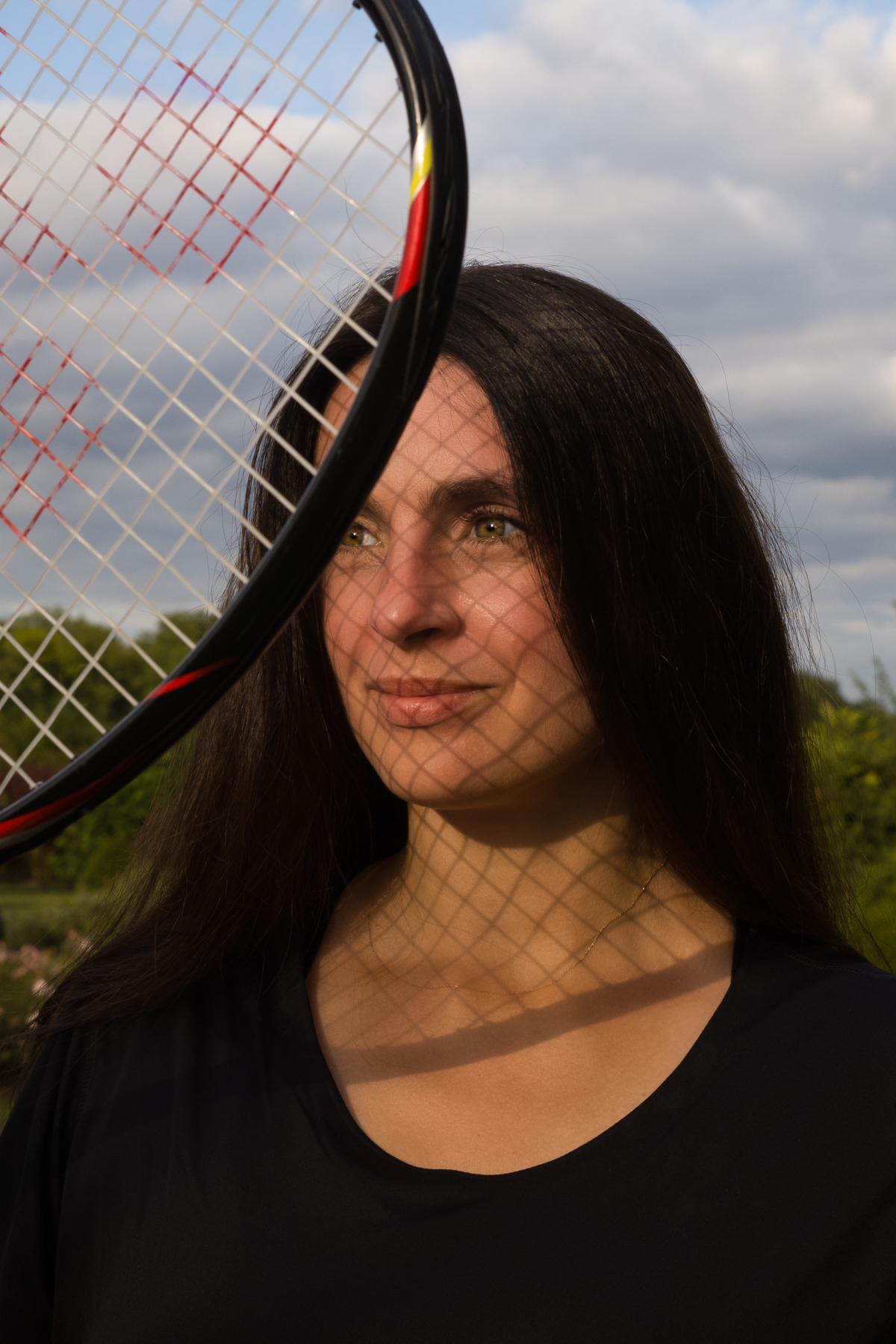 Woman with long dark hair holding a tennis racket, her face framed by the racket's strings, outdoors against cloudy sky