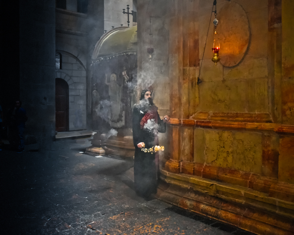 A figure in dark robes stands in a dimly lit stone corridor, surrounded by incense smoke and religious iconography, including a hanging lamp.
