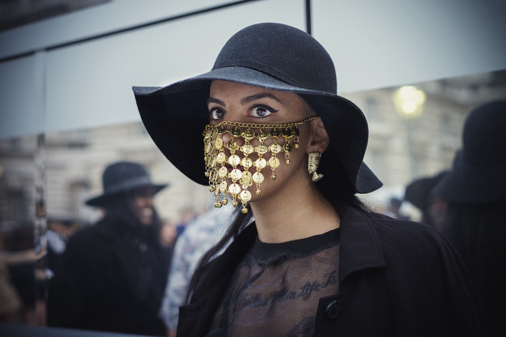 Person wearing wide-brimmed black hat and gold chain mail face covering, looking directly at camera with dramatic eye makeup