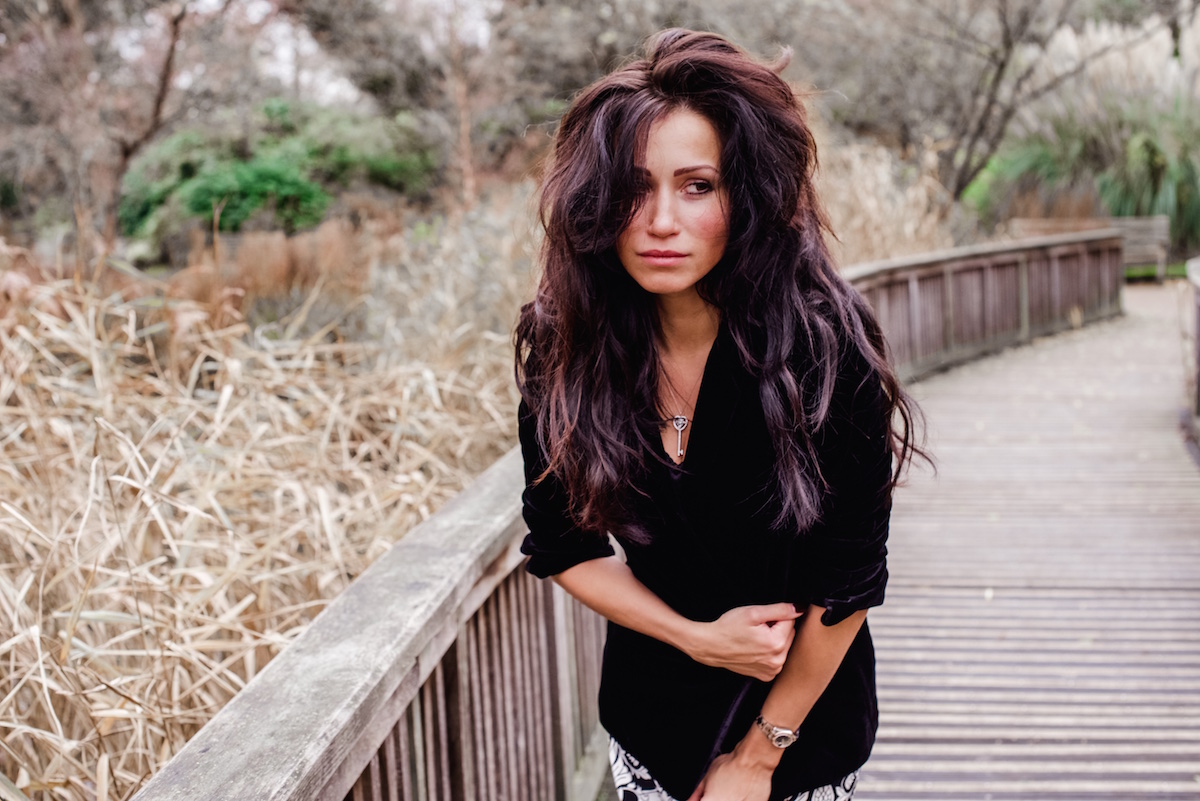 Woman with long dark hair in black clothing standing on a wooden boardwalk, gazing thoughtfully into the distance amid bare trees and winter landscape.