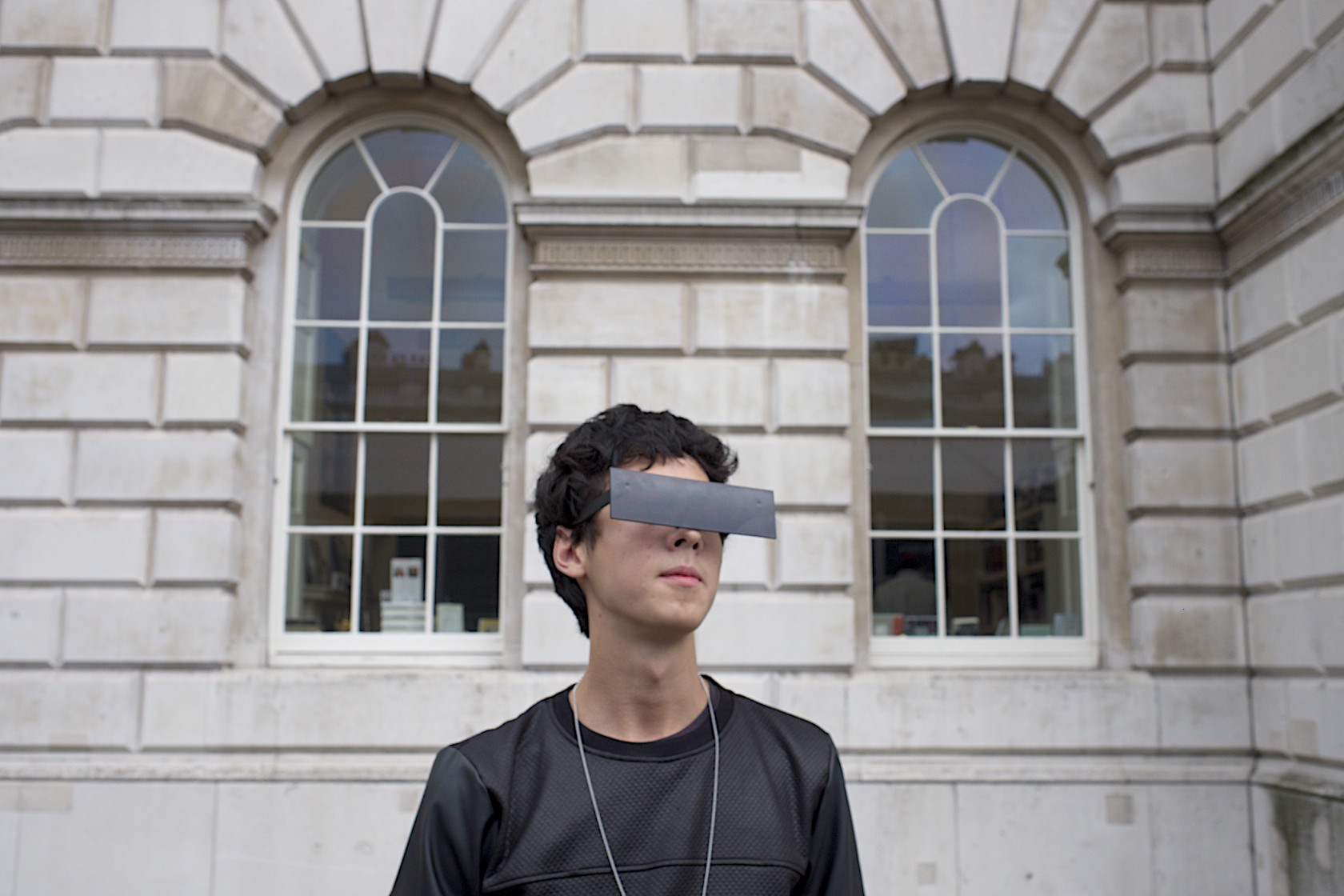 Young person wearing a dark band across their eyes, standing against a classical stone building with large arched windows